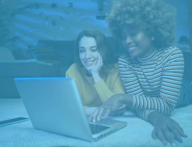 Young multiethnic lesbian couple lying down in the living room at home using laptop searching for weddings venues together during night time.